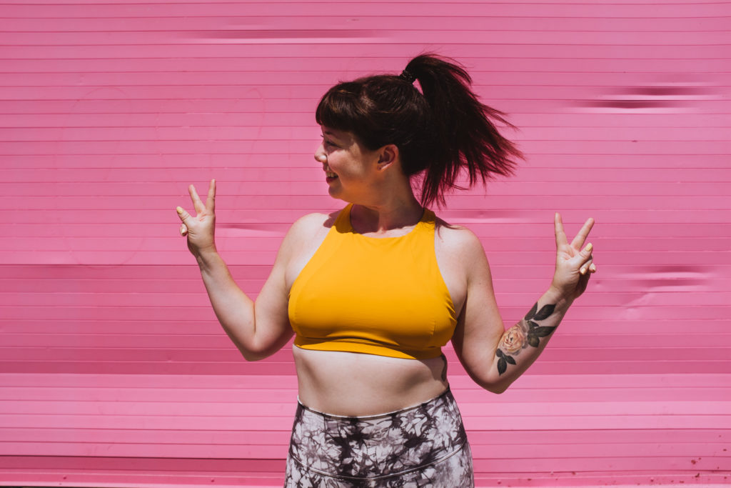 yoga teacher in yellow tank against bright pink wall with peace sign fingers