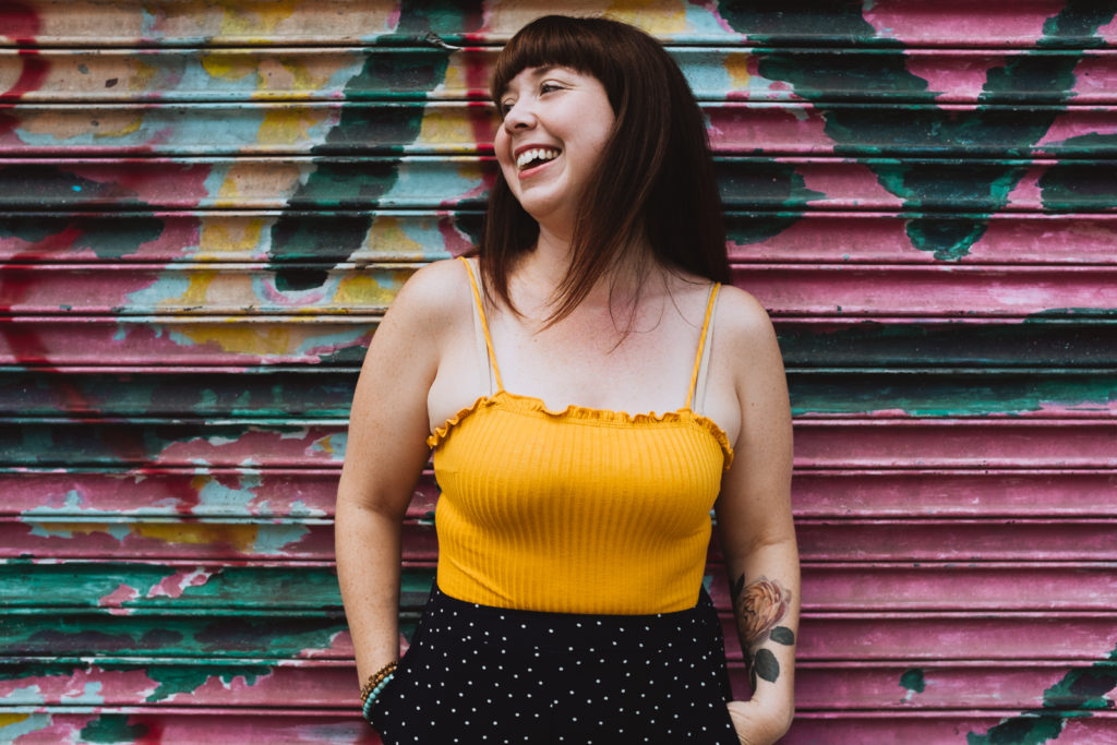 lifestyle portrait of Megan in bright yellow tank laughing against graffiti wall