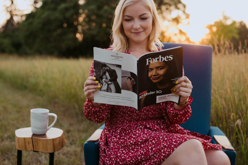 blonde girl reading forbes magazine sitting in a blue chair in a field at sunset