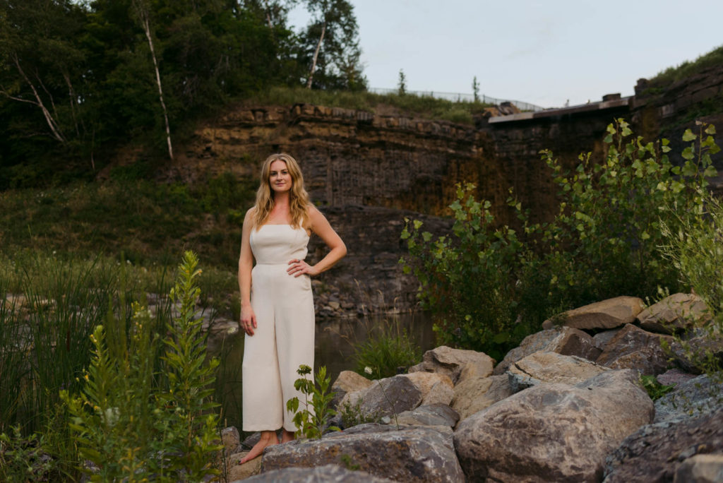 Stephanie Karlovits female entrepreneur wearing a white jumper standing on rocks by a dam
