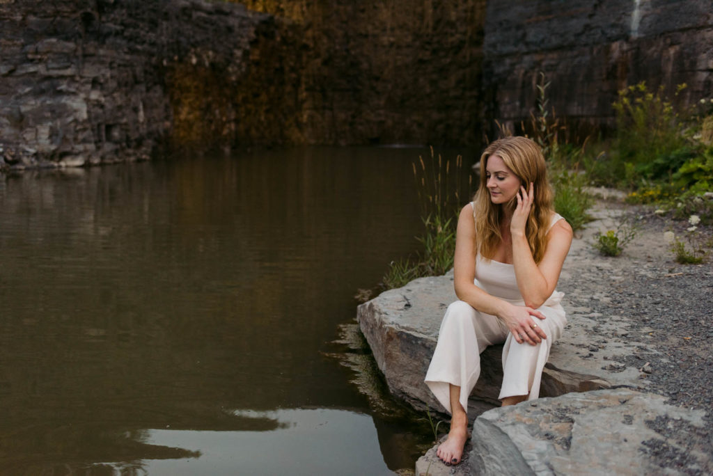 Stephanie Karlovits female entrepreneur wearing a white jumper sitting on rocks by a dam