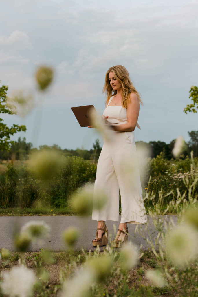 Stephanie Karlovits female entrepreneur wearing a white jumper and holding her laptop outdoors in the tall grass