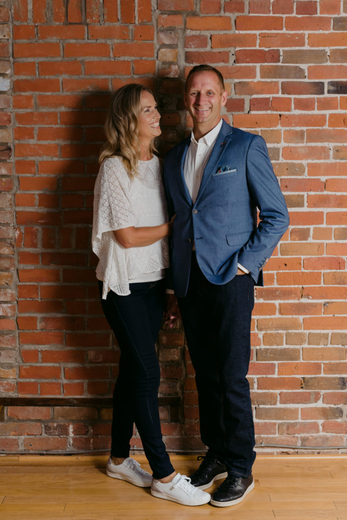 man and wife business owners standing against brick wall smiling at each other