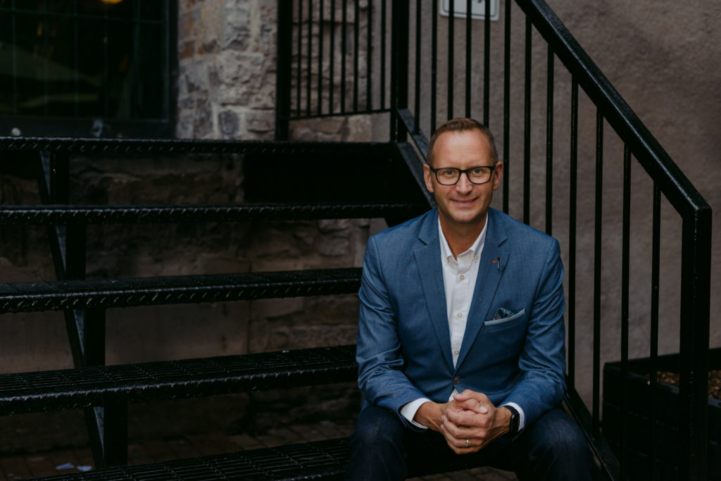 male business owner sitting on black iron steps in a blue suit jacket and glasses