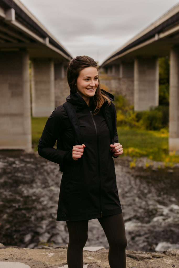 physiotherapist in lululemon rain jacket standing under a bridge by the water with backpack on