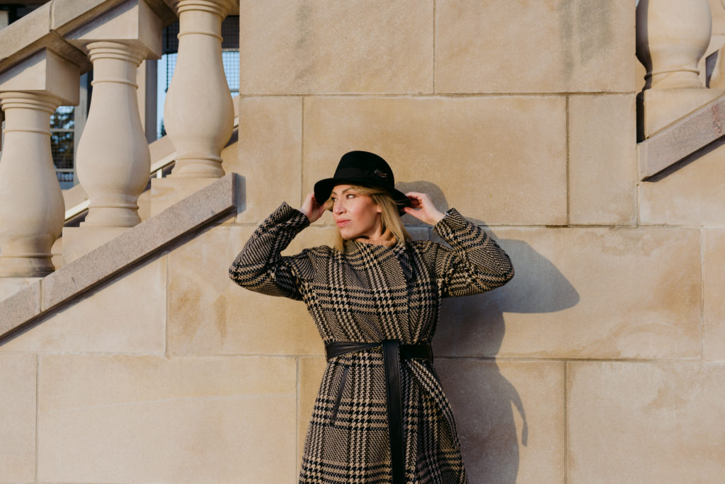 Woman wearing black hat and plaid wool jacket by stone railing at sunset