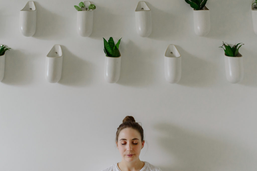 yoga teacher meditating below plants in white pots hanging on the wall