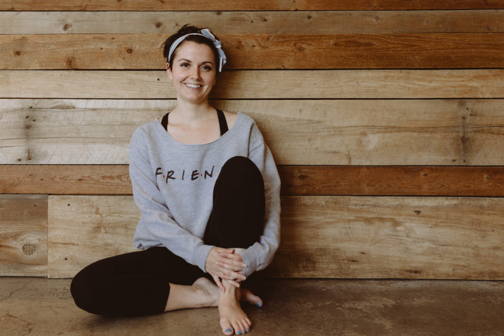 yoga teacher wearing a Friends sweatshirt sitting against barn board wall smiling