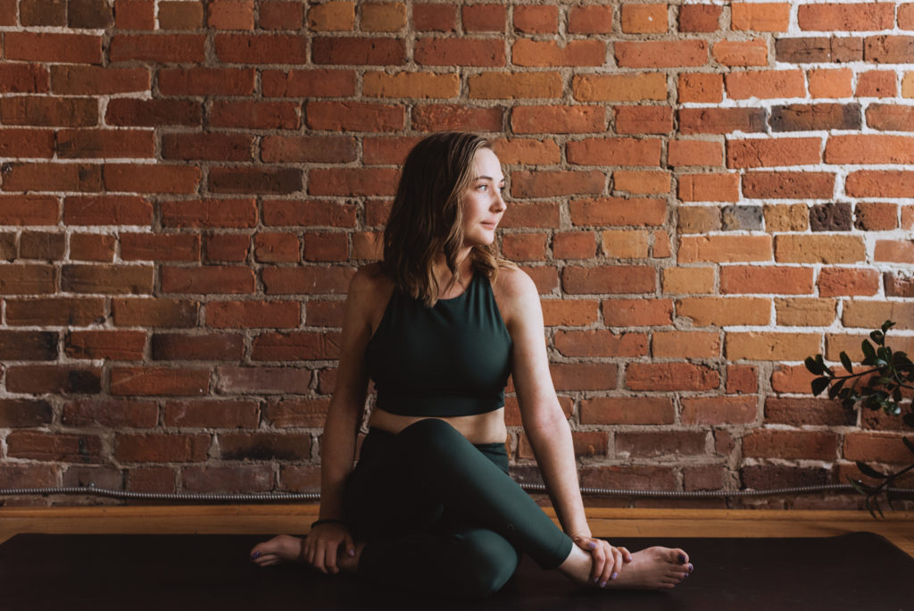 yoga teacher sitting against red brick wall in cow face legs