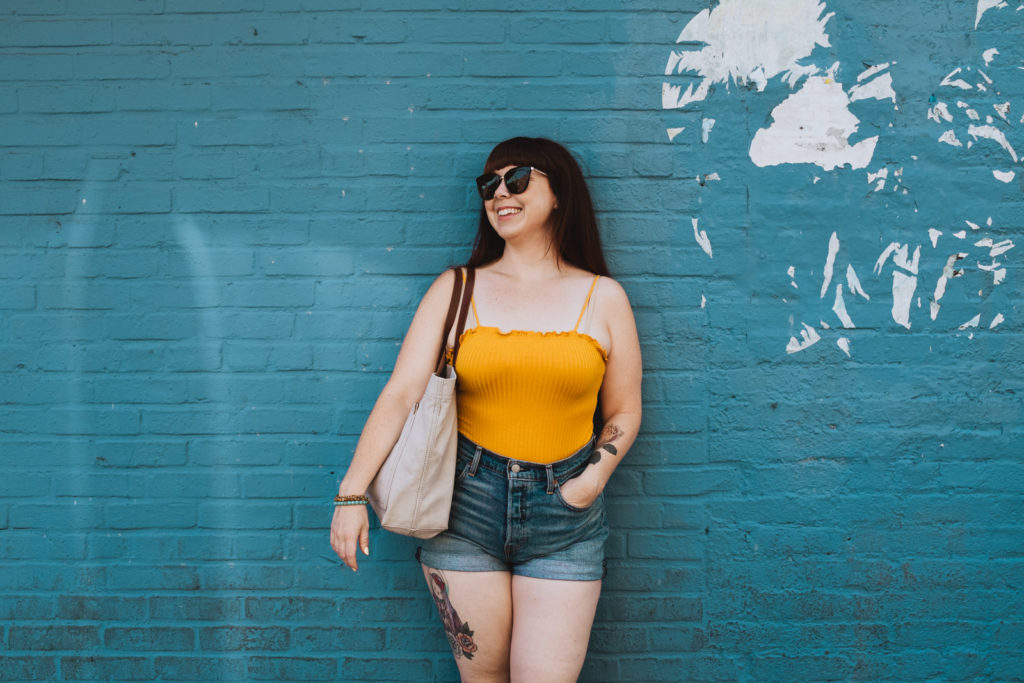 lifestyle photo of Megan wearing jean shorts and bright yellow tank against blue wall