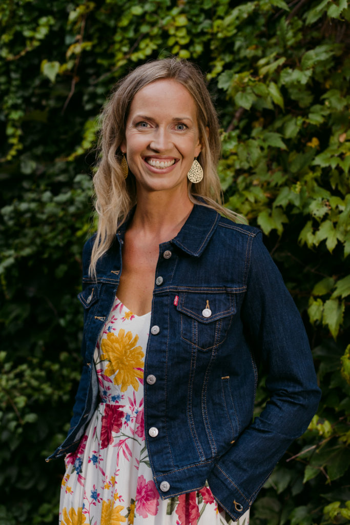 female business owner wearing jean jacket and floral dress in front of vines